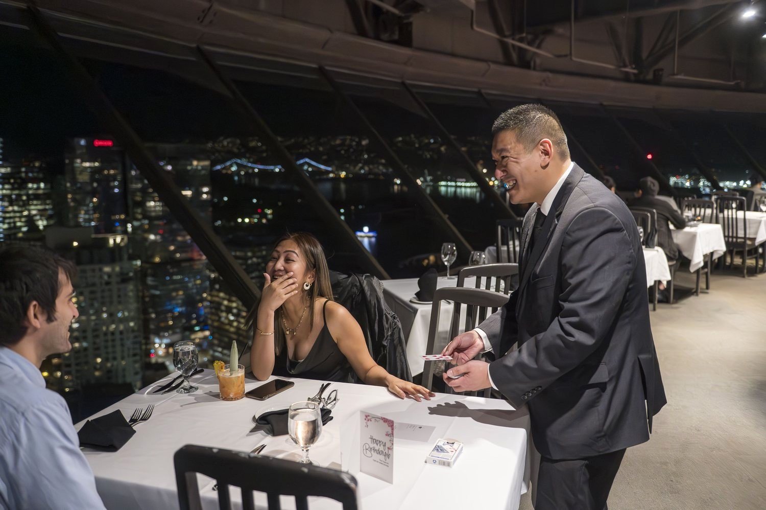 John Ha performing close-up magic for a laughing guest at a Vancouver rooftop event with the city skyline behind them
